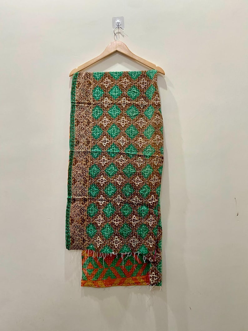 May include: A patterned textile hanging on a wooden hanger. The textile features a repeating diamond pattern in shades of green, brown, and white. The bottom edge has an orange and green geometric design. The textile is likely a blanket or throw.