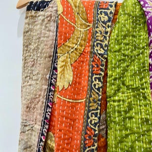 May include: A colorful patchwork textile hanging on a wooden hanger. The textile features various patterned fabrics in shades of orange, green, beige, and purple. The fabrics are stitched together with white thread, creating a textured effect.