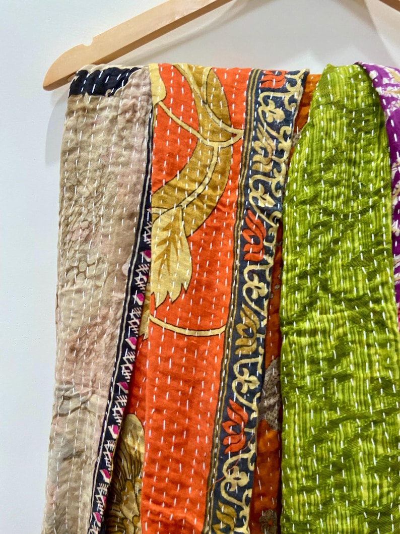 May include: A colorful patchwork textile hanging on a wooden hanger. The textile features various patterned fabrics in shades of orange, green, beige, and purple. The fabrics are stitched together with white thread, creating a textured effect.