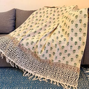 May include: Cream-coloured throw blanket with a repeating green floral pattern. The border features a geometric black and white design with a brown accent and fringe. The blanket is draped over a grey sofa.