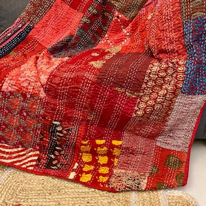 Peut inclure: Une courtepointe patchwork rouge avec divers motifs et textures. La courtepointe est faite de différents tissus et cousue avec du fil blanc. La courtepointe est drapée sur une surface grise.