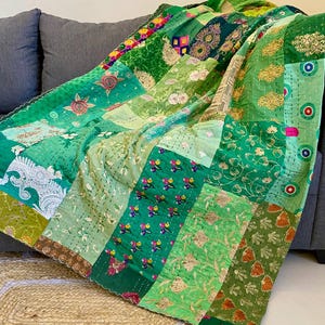 Könnte beinhalten: Ein Patchwork-Quilt in Grüntönen mit floralen und geometrischen Mustern. Der Quilt ist über ein graues Sofa drapiert und zeigt eine Vielzahl von Texturen und Designs. Die Kanten des Quilts sind gewellt, was einen dekorativen Touch verleiht.