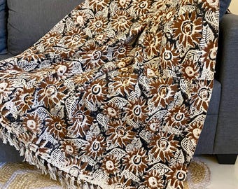 May include: A brown, black, and white floral patterned throw blanket with a fringed edge is draped over a gray sofa. The blanket features a repeating pattern of stylized flowers and leaves. A natural fiber rug is visible below.