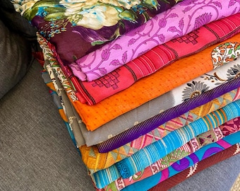 May include: A stack of colorful fabric pieces, showcasing a variety of patterns and colors. The top layer features a floral design with green, blue, and white hues on a dark purple background. Other fabrics display stripes, geometric shapes, and vibrant shades of pink, orange, and blue.