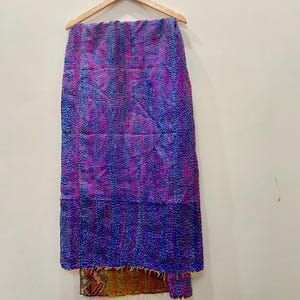Op de afbeelding: Handgemaakt textiel, hangend aan een houten hanger. De stof heeft een patchwork-ontwerp met tinten paars, blauw en roze. De onderste rand heeft een contrasterende stof met kleurrijke franjes.