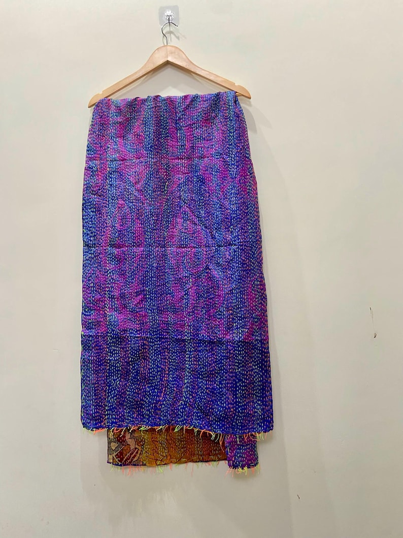 Op de afbeelding: Handgemaakt textiel, hangend aan een houten hanger. De stof heeft een patchwork-ontwerp met tinten paars, blauw en roze. De onderste rand heeft een contrasterende stof met kleurrijke franjes.