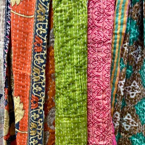 May include: A close-up of colorful fabric strips with various patterns and textures. The strips include orange with floral designs, green with a textured pattern, and pink with a repeating leaf pattern. White stitching is visible throughout.