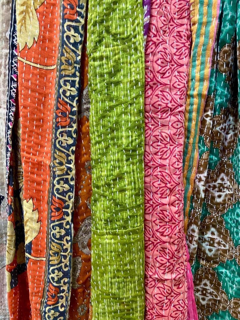 May include: A close-up of colorful fabric strips with various patterns and textures. The strips include orange with floral designs, green with a textured pattern, and pink with a repeating leaf pattern. White stitching is visible throughout.