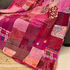 Op de afbeelding: Een patchworkquilt in roze en magenta tinten. De quilt is gemaakt van verschillende stoffen vierkanten met verschillende patronen en texturen, waaronder bloemen- en geometrische ontwerpen. De randen zijn afgewerkt met een roze rand.