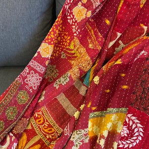 Peut inclure: Textile patchwork aux tons de rouge, jaune et orange. Le tissu présente des motifs et des dessins complexes, notamment des motifs floraux et géométriques. Le textile est probablement une couverture ou un plaid, avec des coutures visibles.