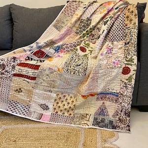 May include: A patchwork quilt draped over a gray sofa. The quilt features a variety of patterned fabric squares in shades of beige, pink, purple, and green. The quilt is resting on a natural fiber rug with a scalloped edge.