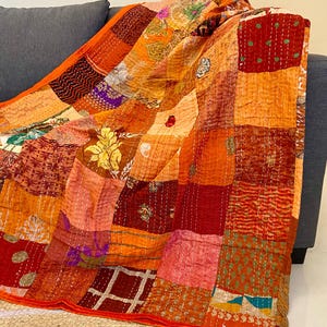 May include: A patchwork quilt in shades of orange, red, and peach, draped over a gray sofa. The quilt features a variety of patterned fabrics, including floral and geometric designs, stitched together with visible seams. The quilt's edges are trimmed with a red border.