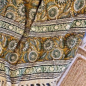 Könnte beinhalten: Senfgelbes und grünes Textil mit Blumenmuster, cremefarbenem Rand und Fransen. Das Textil weist ein sich wiederholendes Muster aus großen Blumen und Blättern in Grün- und Weißtönen auf. Der Rand hat ein dekoratives Design mit schwarzen Linien.