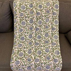 May include: A white fabric with a repeating pattern of blue floral clusters and green leaves on brown stems. The fabric is draped over a gray sofa, showcasing the design and texture of the textile.