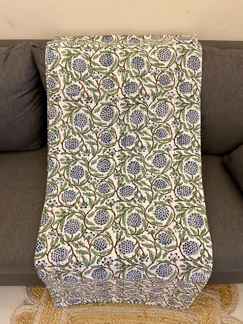 May include: A white fabric with a repeating pattern of blue floral clusters and green leaves on brown stems. The fabric is draped over a gray sofa, showcasing the design and texture of the textile.