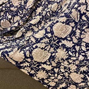 May include: A navy blue and cream floral patterned quilt. The quilt features a repeating design of large flowers and leaves. The fabric is draped over a gray surface, showcasing the intricate stitching and detailed pattern. The quilt is a home decor item.