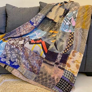 Op de afbeelding: Een patchwork quilt gedrapeerd over een grijze bank. De quilt heeft een verscheidenheid aan stofpatronen en kleuren, waaronder blauw, oranje, beige en bloemmotieven. De quilt is gemaakt van meerdere aan elkaar genaaide stofvierkanten.