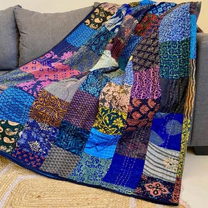 May include: A patchwork quilt with a variety of patterned fabric squares in shades of blue, pink, yellow, and green. The quilt is draped over a gray sofa, showcasing its vibrant colors and textures. The quilt's edges are finished with a dark blue border.