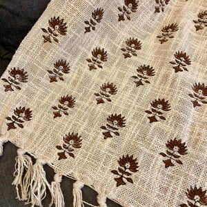 Può includere: Tessuto tessuto color crema con un motivo ripetuto di disegni floreali marroni. Il tessuto ha un bordo sfrangiato con lunghe nappe bianche sporche. Il tessuto è probabilmente un plaid o una sciarpa decorativa.