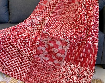 Op de afbeelding: Een rood-wit patroon plaid over een grijze bank. De deken heeft verschillende ontwerpen, waaronder stippen, wervelingen en bloempatronen. De plaid is gemaakt van een zacht materiaal en voegt een vleugje kleur toe aan de kamer.
