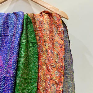 Op de afbeelding: Een houten kleerhanger toont verschillende kleurrijke, bedrukte textielsoorten. De stoffen omvatten een levendig blauw, een diep groen en een oranje met een paisley-ontwerp. Dit zijn waarschijnlijk sjaals of omslagdoeken.