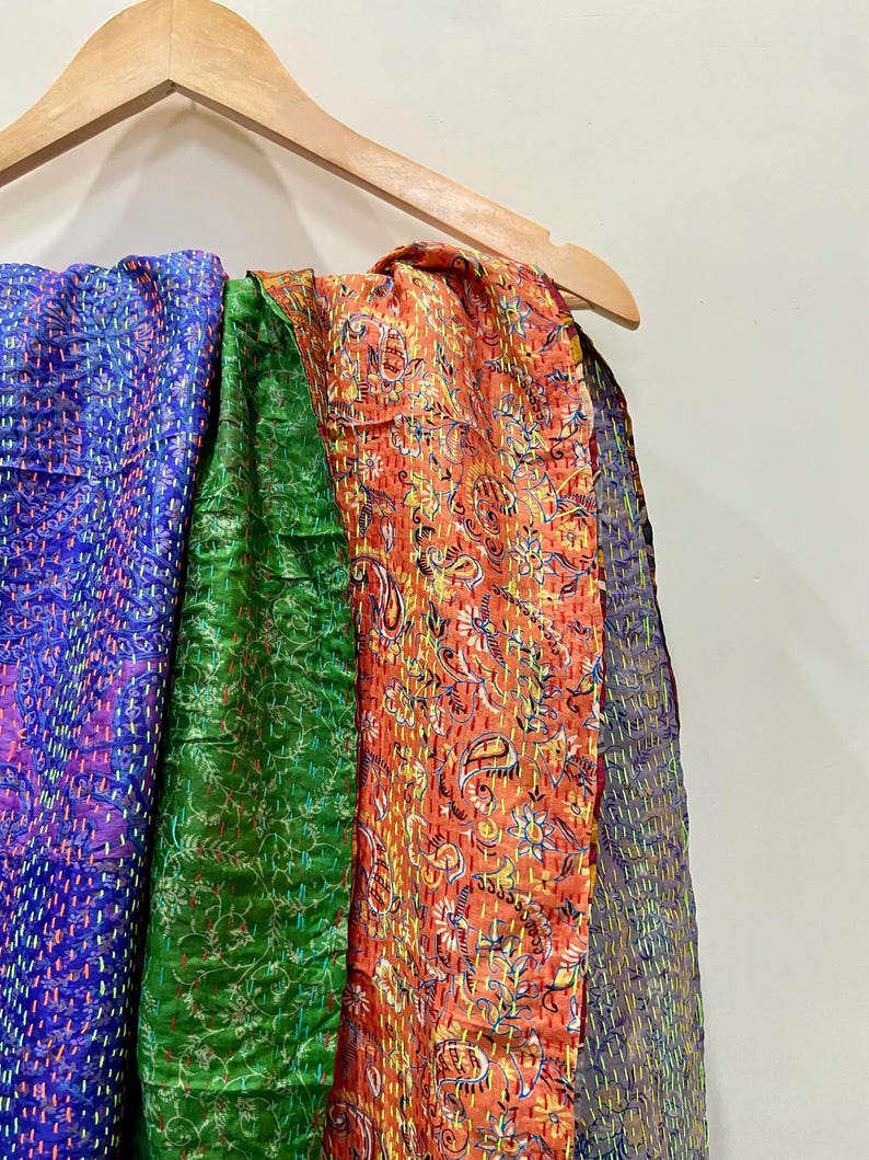 Op de afbeelding: Een houten kleerhanger toont verschillende kleurrijke, bedrukte textielsoorten. De stoffen omvatten een levendig blauw, een diep groen en een oranje met een paisley-ontwerp. Dit zijn waarschijnlijk sjaals of omslagdoeken.