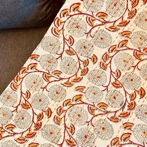 Op de afbeelding: Crèmekleurig textiel met een herhalend bloemmotief in bruin en oranje. Het ontwerp toont grote, gestileerde bloemen en kronkelende wijnranken. De stof lijkt gewatteerd met zichtbare stiksels.