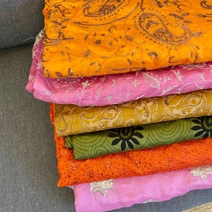 Puede incluir: Una pila de coloridas piezas de tela en tonos naranja, rosa, verde y amarillo. Los tejidos presentan patrones detallados, incluyendo diseños de cachemira y florales, con adornos brillantes. Las telas parecen ser de seda o un material ligero similar.