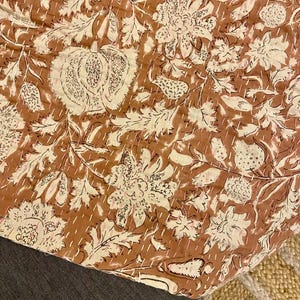 Op de afbeelding: Een bruin en cr&egrave;mekleurig textiel met bloemenpatroon. Het ontwerp toont grote bloemen, bladeren en andere botanische elementen. De stof is gestikt met witte draad, waardoor een gewatteerd effect ontstaat. Het textiel is over een donkergrijze ondergrond gevouwen.