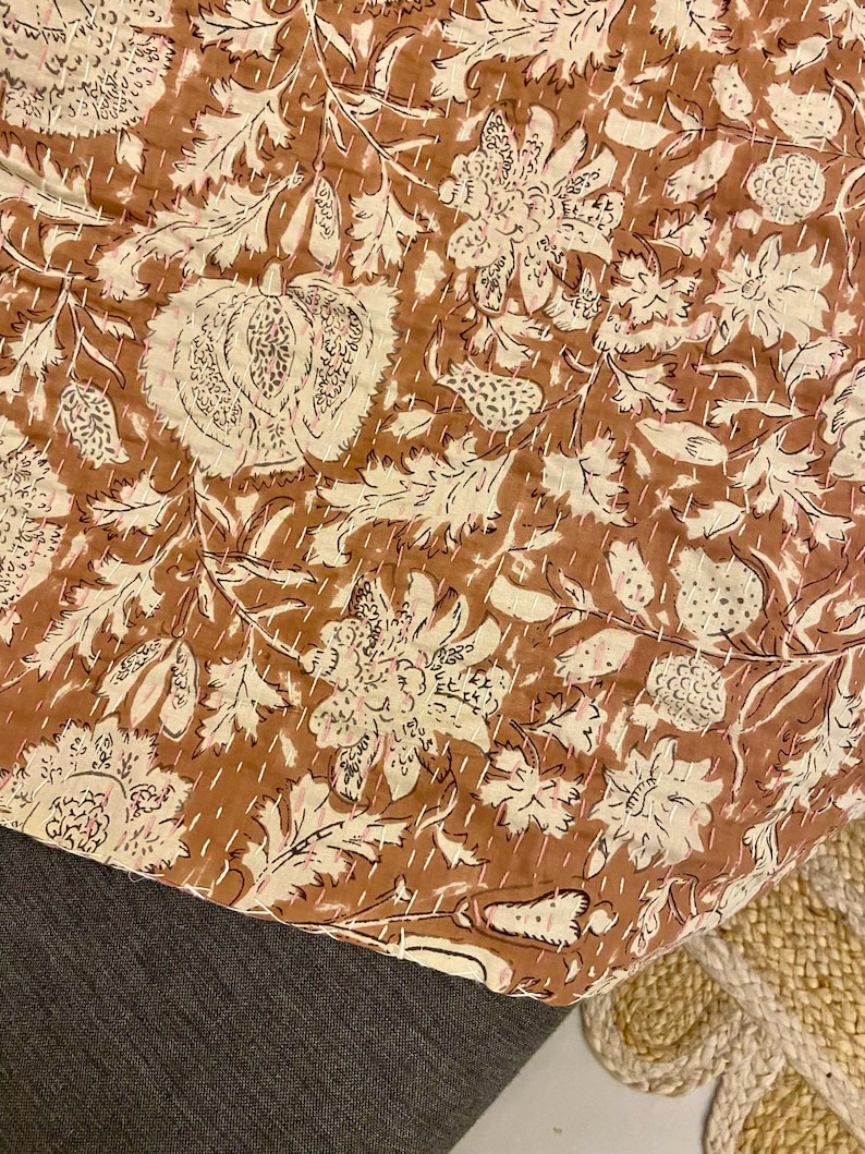 Op de afbeelding: Een bruin en cr&egrave;mekleurig textiel met bloemenpatroon. Het ontwerp toont grote bloemen, bladeren en andere botanische elementen. De stof is gestikt met witte draad, waardoor een gewatteerd effect ontstaat. Het textiel is over een donkergrijze ondergrond gevouwen.