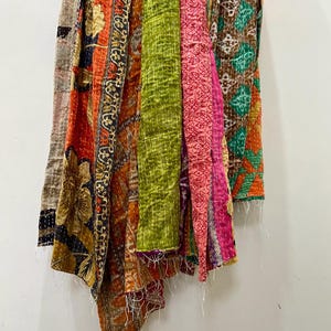 May include: A collection of colorful, patterned fabric pieces hanging against a white wall. The fabrics feature a variety of designs, including floral and geometric patterns, in shades of orange, green, pink, and brown. The edges have visible stitching.