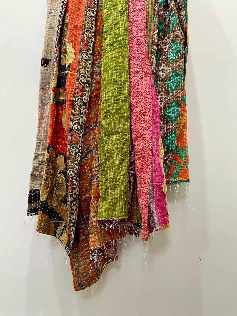 May include: A collection of colorful, patterned fabric pieces hanging against a white wall. The fabrics feature a variety of designs, including floral and geometric patterns, in shades of orange, green, pink, and brown. The edges have visible stitching.