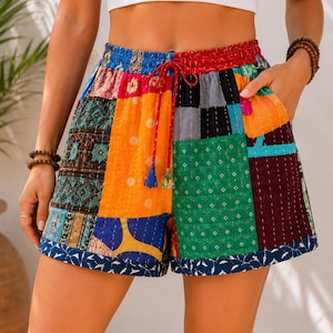 Puede incluir: Pantalones cortos de patchwork con una mezcla vibrante de cuadrados de tela multicolores con detalles de costura blanca visibles. Los pantalones tienen una cintura elástica con cordón rojo, bolsillos laterales y un dobladillo azul oscuro.