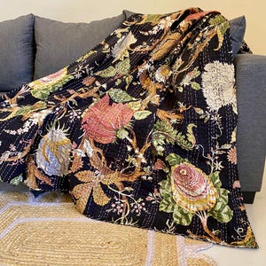 Pode incluir: Um edredom floral preto drapeado sobre um sofá cinza. O edredom apresenta um padrão vibrante de grandes flores em tons de rosa, verde e amarelo, com detalhes de costura intrincados. O edredom repousa sobre um tapete de fibra natural.