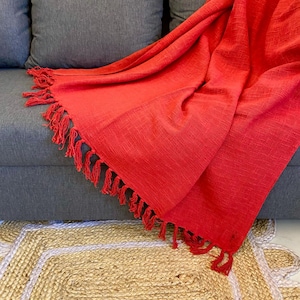 Könnte beinhalten: Eine leuchtend rote Decke mit Fransen, die über einem grauen Sofa drapiert ist. Die Textur der Decke ist sichtbar und kontrastiert mit dem Sofa und einem Jute-Teppich im Vordergrund. Die Decke sorgt für einen Farbtupfer.