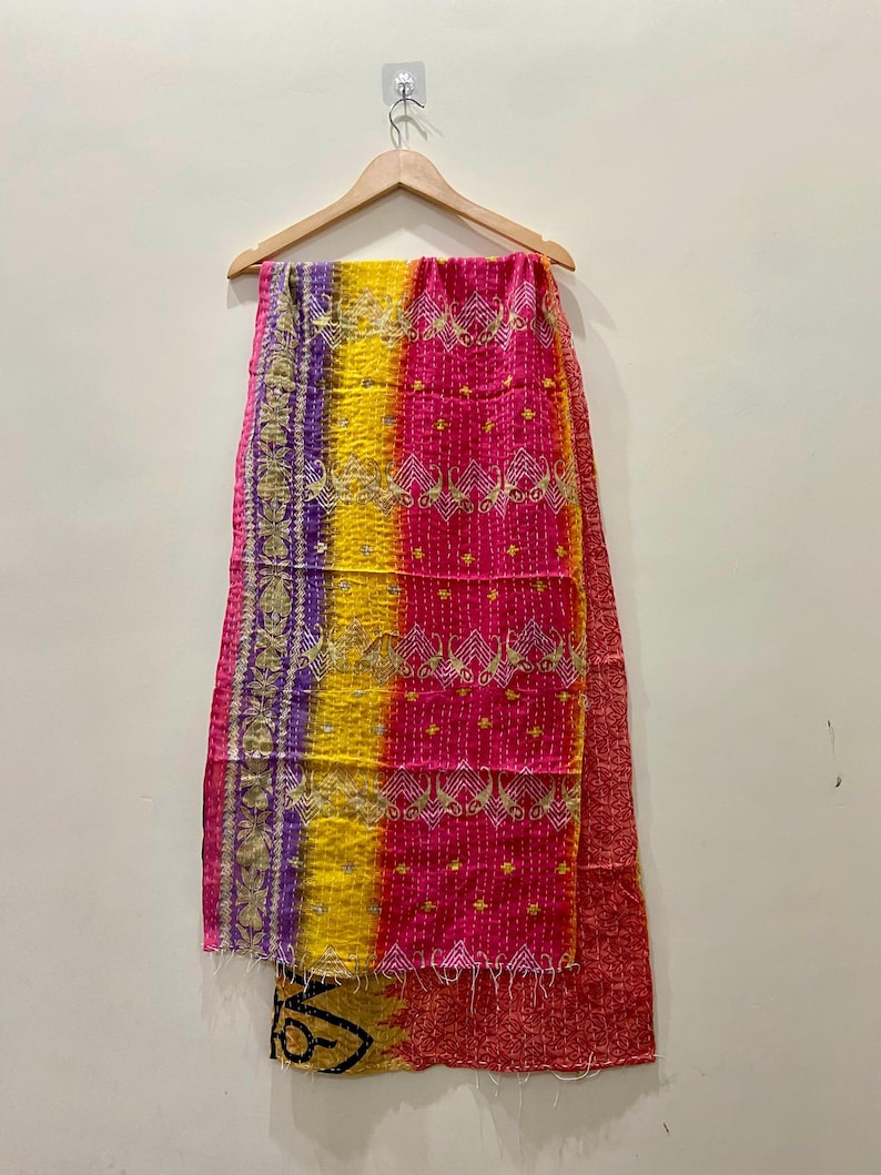 May include: A colorful textile hanging on a wooden hanger. The textile features vertical stripes of purple, yellow, and pink, with intricate embroidered patterns and a red border. The fabric appears to be made of a soft material.