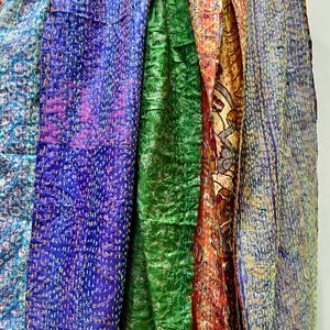 Op de afbeelding: Een verzameling kleurrijke, bedrukte sjaals die aan een houten hanger hangen. De sjaals tonen een verscheidenheid aan kleuren, waaronder blauw, paars, groen, oranje en grijs. Elke sjaal heeft een uniek ontwerp en textuur, met zichtbare stiksels.