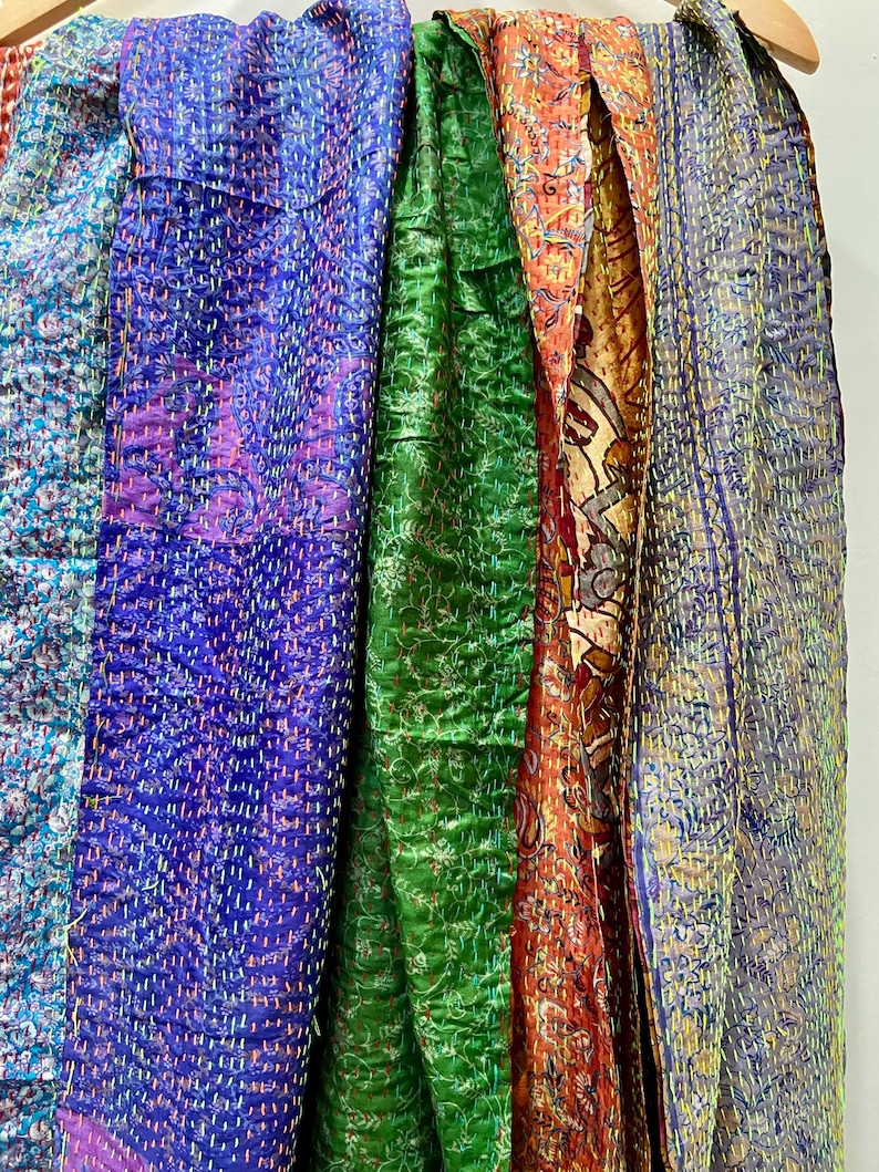 Op de afbeelding: Een verzameling kleurrijke, bedrukte sjaals die aan een houten hanger hangen. De sjaals tonen een verscheidenheid aan kleuren, waaronder blauw, paars, groen, oranje en grijs. Elke sjaal heeft een uniek ontwerp en textuur, met zichtbare stiksels.