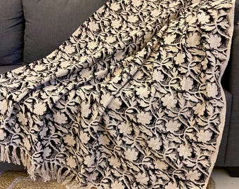 May include: A black and white floral patterned throw blanket with fringe detail draped over a gray sofa. The blanket's design features a repeating floral motif. The throw is made of a soft, textured fabric, perfect for adding a touch of style to any living space.