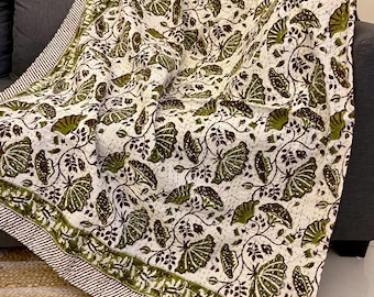 May include: A white and green patterned quilt draped over a gray sofa. The quilt features a repeating design of green leaves and brown vines. The border is a striped pattern in brown and white.