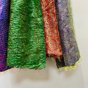 Op de afbeelding: Close-up van kleurrijke, getextureerde stoffen panelen. De panelen zijn in tinten paars, groen, rood en blauw. Elk paneel heeft een uniek patroon en is gestikt met contrasterend garen, waardoor een patchwork-effect ontstaat. De randen hebben losse draden.