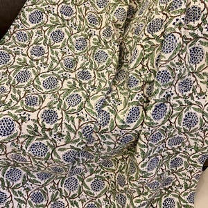 May include: A white fabric with a repeating pattern of blue and green floral designs. The pattern features clusters of blue flowers with green leaves and brown stems. The fabric is draped, showing folds and texture.