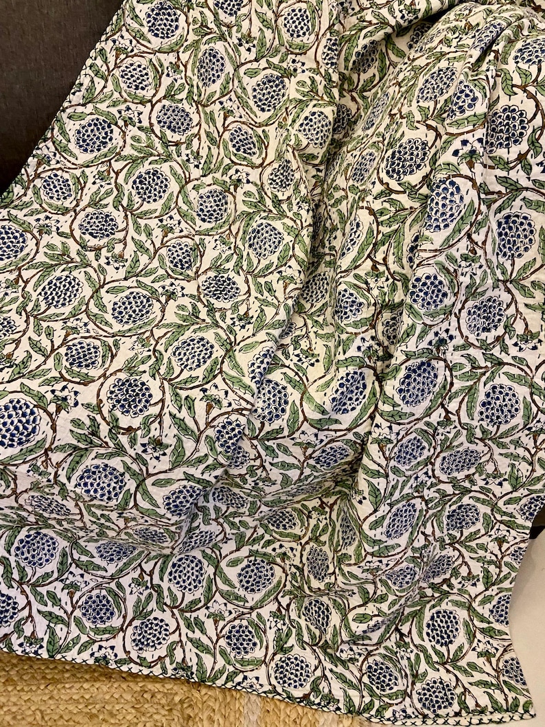 May include: A white fabric with a repeating pattern of blue and green floral designs. The pattern features clusters of blue flowers with green leaves and brown stems. The fabric is draped, showing folds and texture.