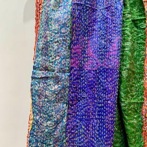 Op de afbeelding: Close-up van kleurrijke, gestikte stof. De textielstukken hebben verschillende patronen en kleuren, waaronder teal, paars en groen. De stof hangt aan een houten hanger, wat de ingewikkelde stiksels en het ontwerp benadrukt.