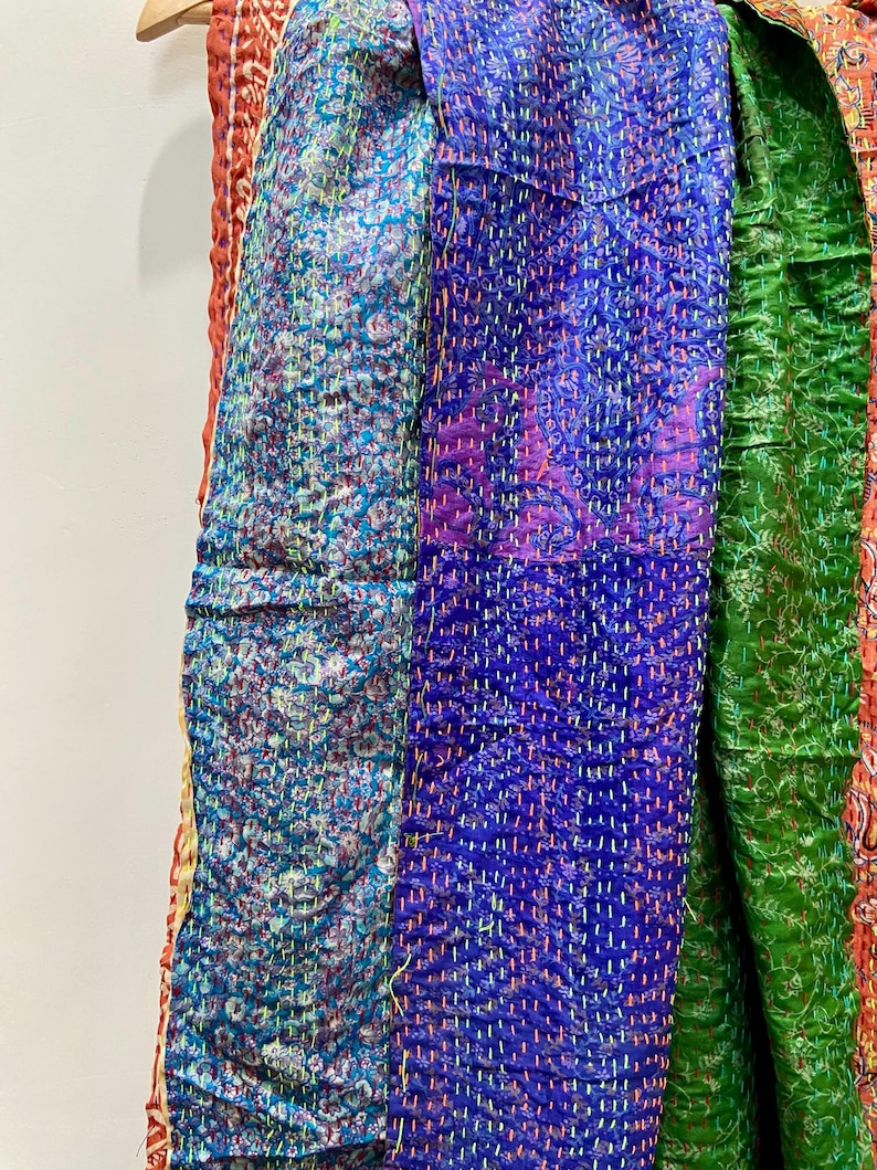 Op de afbeelding: Close-up van kleurrijke, gestikte stof. De textielstukken hebben verschillende patronen en kleuren, waaronder teal, paars en groen. De stof hangt aan een houten hanger, wat de ingewikkelde stiksels en het ontwerp benadrukt.
