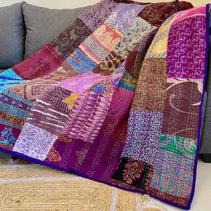 May include: A patchwork quilt in shades of purple, pink, and blue, with a deep purple border. The quilt is draped over a grey sofa, showcasing a variety of patterned fabric squares. The quilt is made of soft material.