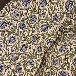 May include: Cream-colored fabric with a repeating pattern of blue floral clusters, green leaves, and brown stems. The design is printed on the fabric, creating a textured appearance. The fabric is folded, showing the pattern's continuity.