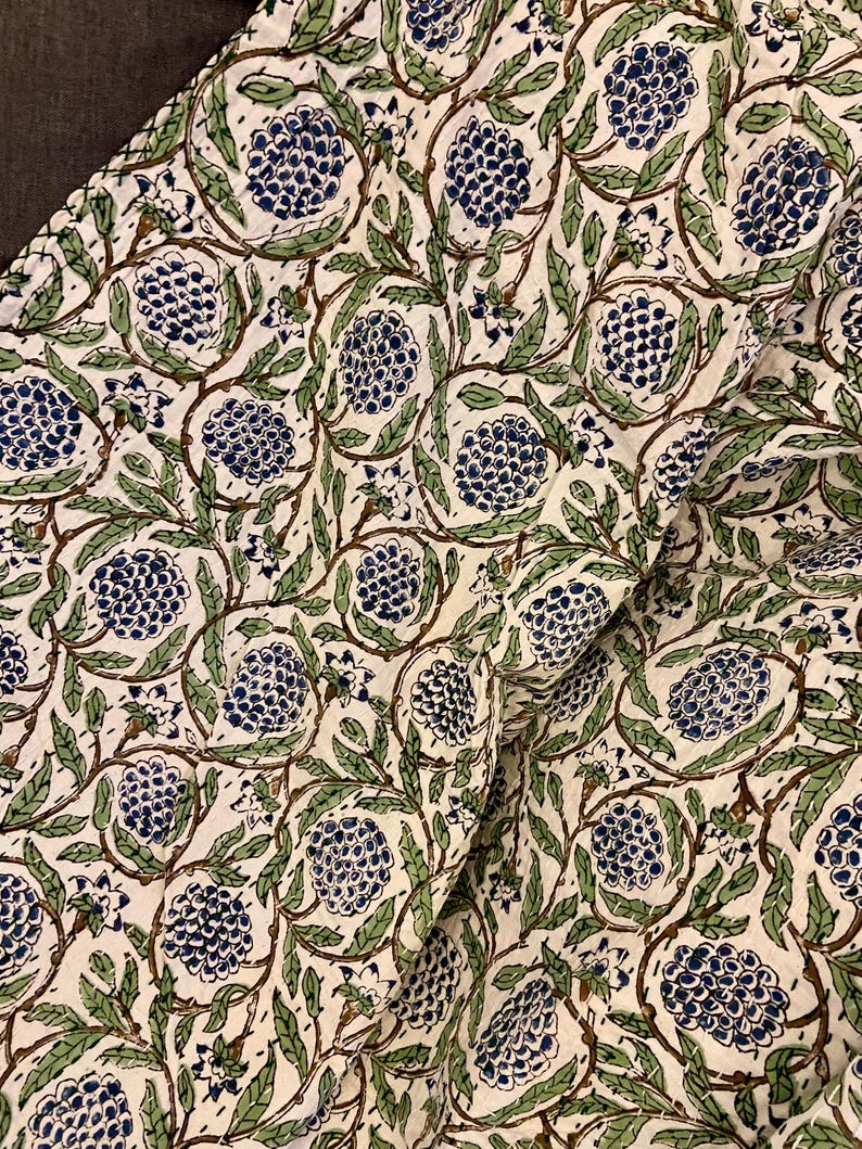 May include: Cream-colored fabric with a repeating pattern of blue floral clusters, green leaves, and brown stems. The design is printed on the fabric, creating a textured appearance. The fabric is folded, showing the pattern's continuity.