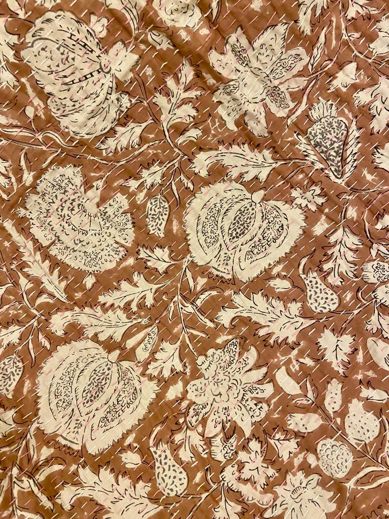 Op de afbeelding: Close-up van een bruin en cr&egrave;mekleurig bloemenpatroon textiel. Het ontwerp toont grote gestileerde bloemen en bladeren met ingewikkelde details. De stof is gestikt met een contrasterend garen, waardoor een gewatteerd effect ontstaat. De algehele esthetiek is vintage en handgemaakt.