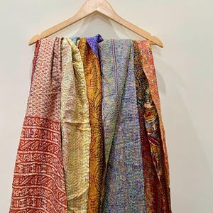 Op de afbeelding: Een verzameling kleurrijke, bedrukte sjaals die aan een houten kleerhanger hangen. De sjaals tonen een verscheidenheid aan kleuren, waaronder rood, geel, blauw en oranje, met ingewikkelde gestikte ontwerpen. De stoffen zijn gedrapeerd en tonen hun texturen en patronen.